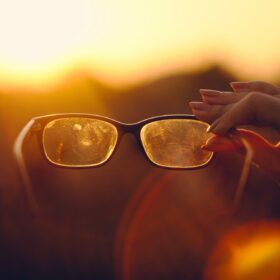 Eine Hand hält eine schutzige Brille gegen den Hintergrund eines Sonnenuntergangs.