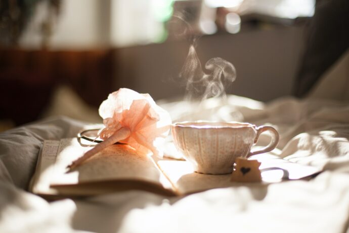Ein Schreibjournal liegt auf einem Bett. Darauf liegt ein rosa Stift mit Blumenbausch. Daneben steht eine Tasse Tee, aus der Dampf emporsteigt.